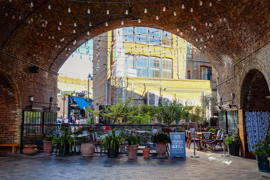 A covered outdoor space beneath a brick arch bridge features a paved flooring area decorated with various potted plants, including lush green foliage and small trees. String lights hang overhead, crea